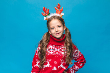 Cute little girl wearing a red Christmas sweater decorated with reindeer and a headband with reindeer antlers smiles and poses against a blue background. Santa Claus helper concept. High quality photo