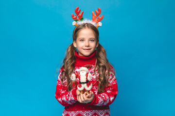 Cute little girl wearing a red Christmas sweater decorated with reindeer and a headband with reindeer antlers holds Santa Claus toy on blue background. High quality photo