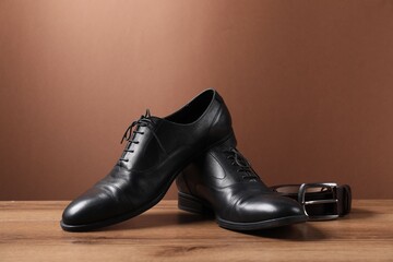 Pair of black leather shoes and belt on wooden surface against brown background