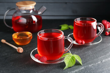 Aromatic raspberry tea in glass cups, teapot, berries, green leaves and honey on black table,...