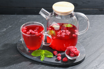 Aromatic raspberry tea in glass cup, teapot, berries and mint on black table, closeup
