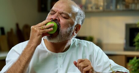 Senior Indian man eating green apple in modern kitchen in morning, Asian old health-conscious man enjoying nutritious fruit for wellness, diet, and natural clean eating lifestyle at home