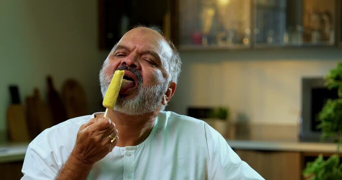 Senior Indian man secretly eating kulfi in modern kitchen, Asian elderly foodie enjoying traditional frozen Indian dessert with guilty pleasure, hiding while savoring sweet creamy kulfi at home
