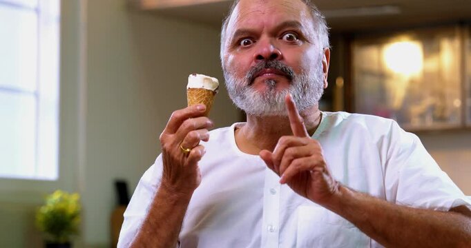 Senior Indian man eating ice cream in cone with delight at home, Asian elderly foodie enjoying cold dessert with childlike joy in modern kitchen, happily savoring frozen creamy treat in summer