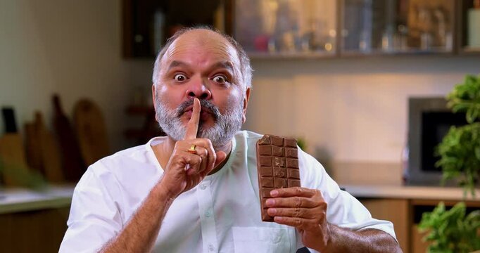 Senior Indian man secretly eating chocolate brick in modern kitchen, Asian elderly foodie sneaking thick chocolate slab with mischievous expression, enjoying sweet treat alone at home