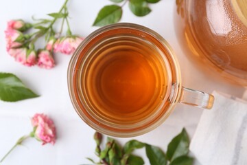 Aromatic tea in glass cup, roses and green leaves on white table, flat lay