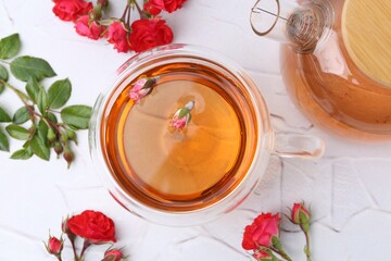 Aromatic tea in glass cup, teapot, roses and green leaves on white table, flat lay