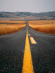 Fototapeta premium Long empty asphalt road stretching into the distance with rolling hills and dry grass fields under a cloudy sky
