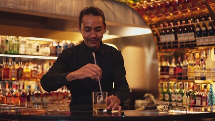 Smart Asian bartender holding stirring mixed alcohol syrup into glass with ice for on the rock...