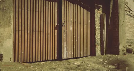 A weathered wooden gate stands closed, surrounded by a textured stone wall. The ground is uneven and littered with debris, suggesting an aged environment. The atmosphere feels quiet. - Powered by Adobe
