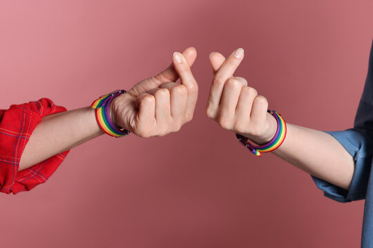 LGBT concept. Women in rainbow wristbands making heart gestures on pink background, closeup
