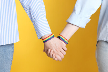 LGBT concept. Women in rainbow wristbands holding hands on yellow background, closeup