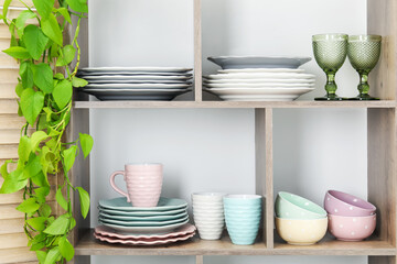 Shelving unit with different dishware, glasses, cups and floral decor near white wall indoors