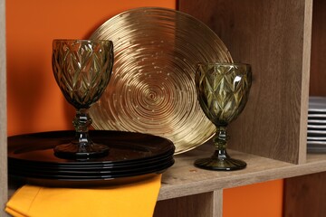 Different dishware and glasses on wooden shelf near orange wall indoors, closeup