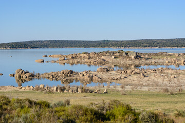 A tranquil lake with clear reflections of rocks and surrounding hills under a bright blue sky. The scene features dry vegetation in the foreground and a peaceful, untouched natural environment, perfec