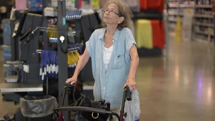 An elderly senior woman navigates through the self checkout area of a hardware store while using a walker. She appears focused on her shopping.
