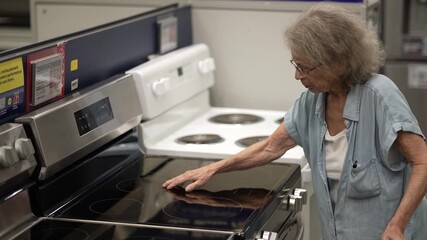 An elderly woman using a walker inspects home remodeling items in a hardware store. She examines a new stove, considering options for her home project.