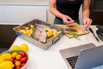 Woman Cooking Snapper Fish with Potatoes and Herbs Using Online Recipe in a Contemporary Kitchen