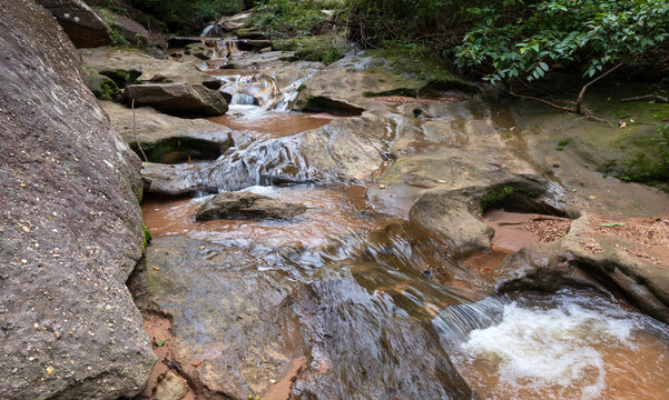 A scenic small stream from Pha Phueng Waterfall winding through mossy rocks in Phu Pha Yon National Park, Thailand, surrounded by green tropical forest during the rainy season.