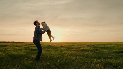Fototapeta premium Dad throws happy daughter up in air with his hands in sun rays. Father, little child play and rejoice together. Happy family weekend in nature. Child in parent arms. Happy family concept. Green field