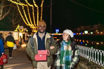 Young multi ethnic couple walking arm in arm carrying christmas gifts at an outdoor night market...