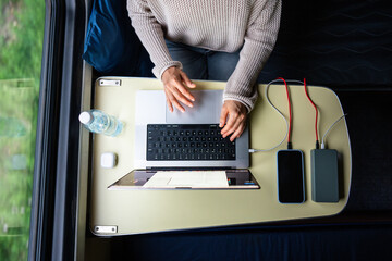 Woman working on laptop to pass the time while traveling. Computer and smartphone connected to modern high-capacity power bank, charging batteries, sitting at table in train compartment, top view.