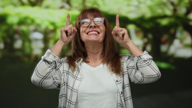 Senior hispanic woman smiling in a vibrant outdoor park setting, pointing upwards with both fingers, conveying happiness and positivity amidst a lush green backdrop