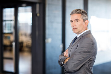 Man in gray suit standing with arms crossed in corridor near glass partition, bright window wall