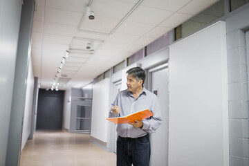 Man inspecting office corridor with paneled walls holding orange folder and pen near ceiling lights