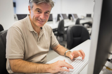 White keyboard resting on desk beside monitor in computer lab, with rows of swivel chairs