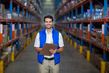 Warehouse worker checking stock levels on shelving racks in aisle, holding clipboard and pen