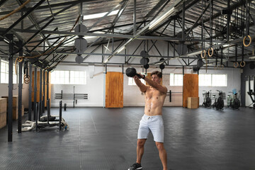 Mid adult man swinging kettlebell on rubber floor in warehouse gym with gymnastic rings and ropes