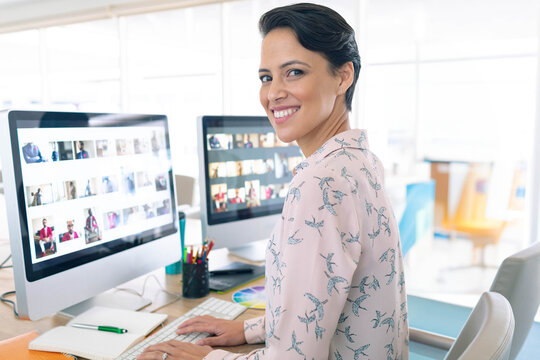 Dual monitors displaying thumbnail grids on wooden desk with color wheel and pencils near window