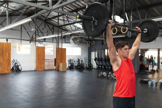 Mid adult man in red sleeveless shirt lifting barbell overhead under gymnastic rings, copy space