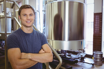 Brewery tech standing arms crossed in brewery next to steel fermentation tank, machinery and kegs