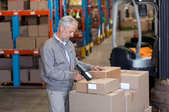 Warehouse worker scanning cardboard boxes with barcode scanner between racks near parked forklift