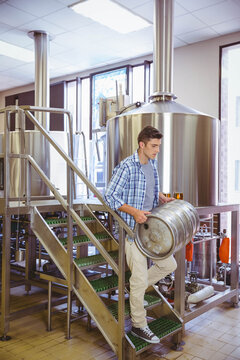 Naklejki Brewery worker descending grated staircase carrying steel beer keg among fermentation tanks