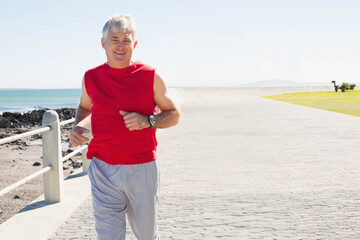 Senior man jogging along paved seaside promenade checking wristwatch near metal railing, copy space
