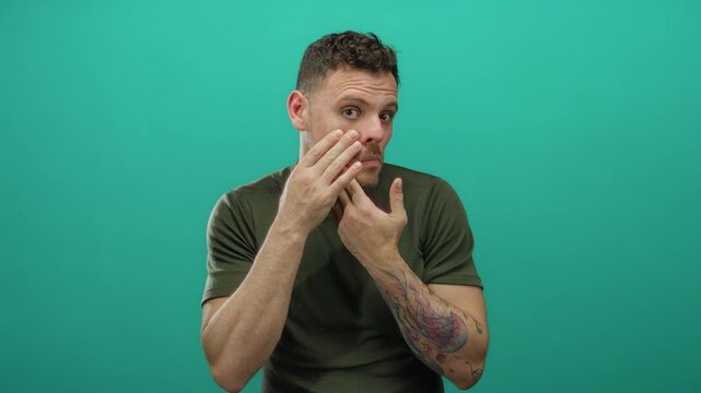 Young man with tattoos touching face against an isolated green background wearing casual clothing with concerned expression showcasing personal care and skincare awareness