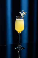 Glass of yellow cocktail with white flowers on a reflective black surface against a dark blue background