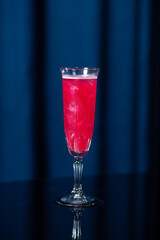 A tall glass filled with a bright pink cocktail with ice cubes, placed on a reflective black...