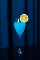 Glass of bright blue cocktail with a lemon slice on the rim, placed on a black reflective surface...