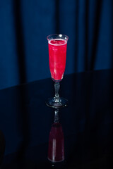 A tall glass filled with a bright pink cocktail with ice cubes, placed on a reflective black surface against a dark blue background