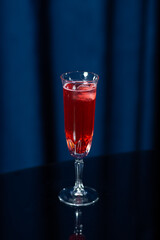 A tall glass filled with a bright red cocktail with ice cubes, placed on a reflective black surface...