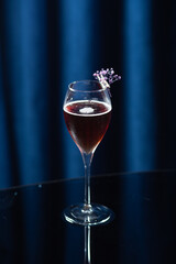 A wine glass filled with a dark red cocktail, decorated with a small purple flower, placed on a reflective black surface with a dark blue background