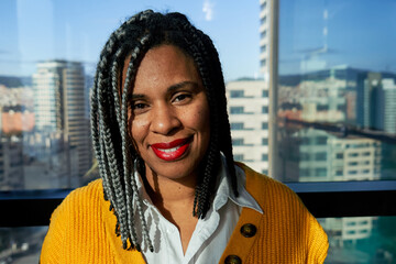 Portrait of an African woman in a modern office with large windows and a city view, looking at camera cheerfully at work.