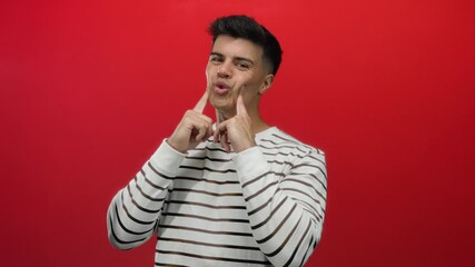 Young man gestures playfully with puffed cheeks wearing striped sweater against vibrant red background implying humorous expression and playful mood in portrait setting.