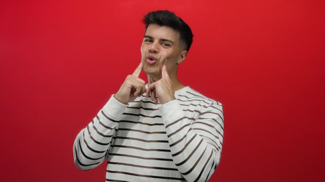 Young man gestures playfully with puffed cheeks wearing striped sweater against vibrant red background implying humorous expression and playful mood in portrait setting.