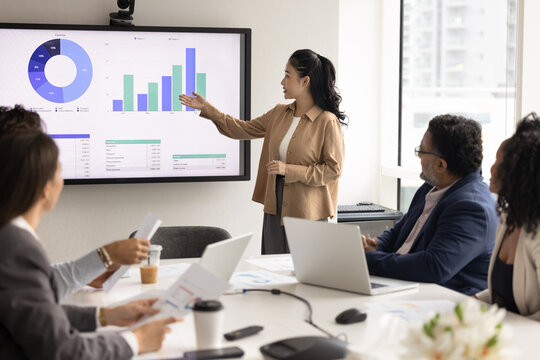 Modern business presentation. Young businesswoman speak to multiethnic professionals group in boardroom meeting explain b2b sales strategy present project stats in charts graphs on digital whiteboard