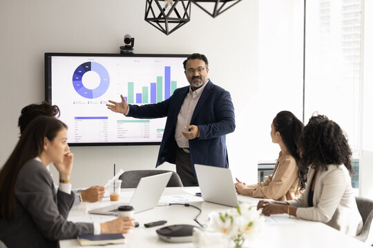 Interactive corporate education workshop. Mature businessman team leader present project statistic data trends on large screen in boardroom to diverse young managers group for collaborative learning
